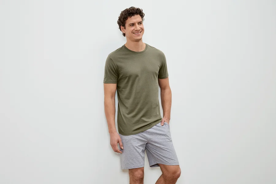 Mann mit lockigem Haar, olivgrünem T-Shirt und grauen Shorts steht vor einer weißen Wand und lächelt.
