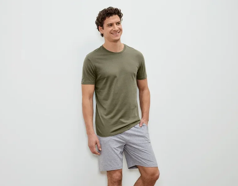 Mann mit lockigem Haar, olivgrünem T-Shirt und grauen Shorts steht vor einer weißen Wand und lächelt.