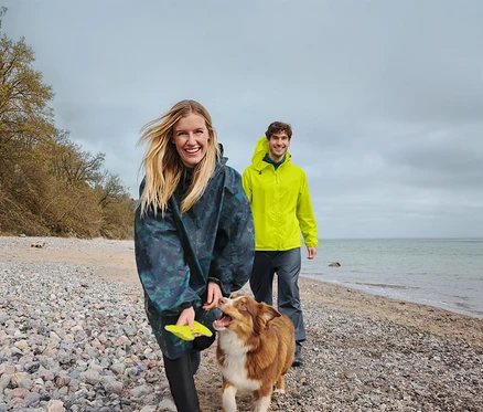 Eine lächelnde Frau in einem dunklen Regenponcho hält einen gelben Frisbee, während sie mit einem braun-weißen Hund an einem Kiesstrand spaziert. Ein lächelnder Mann in einer gelben Regenjacke geht hinter ihnen, mit Meer und bewölktem Himmel im Hintergrund.