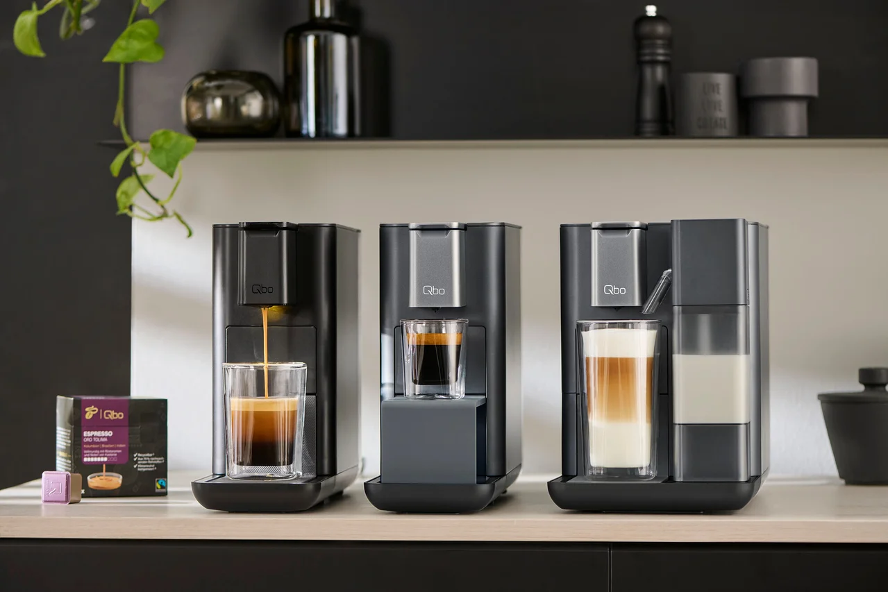 Drei Qbo Kaffeemaschinen stehen auf einer Theke, aus der linken läuft Kaffee in ein Glas. Neben der linken Kaffeemaschine befindet sich eine Packung Qbo Espresso India.