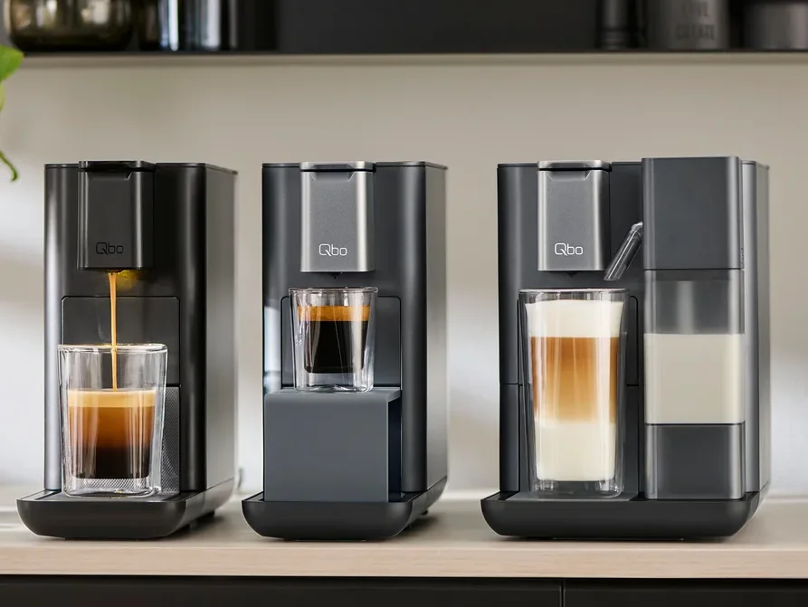Drei Qbo Kaffeemaschinen stehen auf einer Theke, aus der linken läuft Kaffee in ein Glas. Neben der linken Kaffeemaschine befindet sich eine Packung Qbo Espresso India.