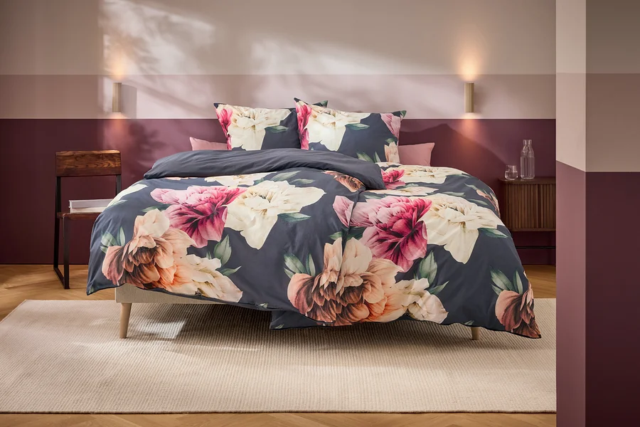 Ein Bett mit dunkelgrauer Bettwäsche und großen rosa, weißen und orangefarbenen Blumenmustern steht in einem Schlafzimmer mit zweifarbiger Wand und Holzmöbeln.