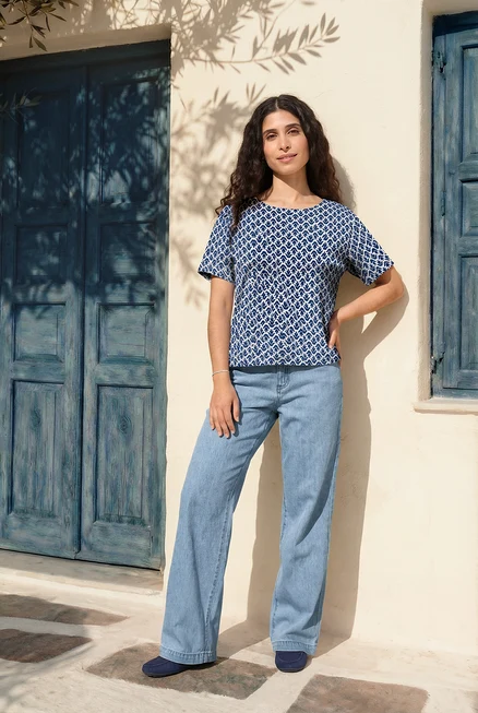 Frau posiert in Blusenshirt mit Alloverprint, Palazzo Jeans, Veloursleder-Loafern und Tennisarmband vor blauem Hauseingang.