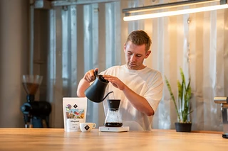 Mann gießt Wasser aus einem schwarzen Kessel in eine Kaffeemaschine. Auf dem Tisch liegen eine Packung Kaffee und eine Tasse.