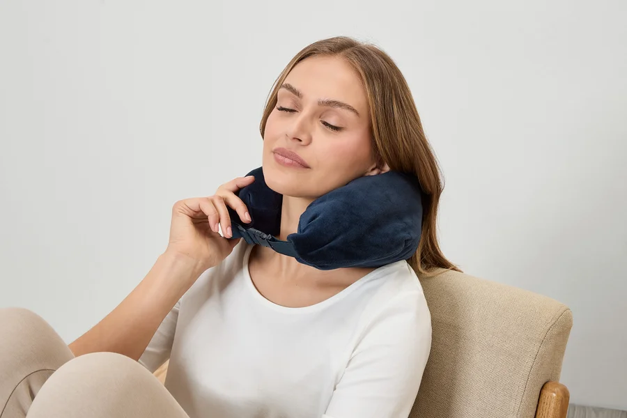 Frau mit geschlossenen Augen trägt eine dunkelblaue Nackenstütze, während sie in einem Sessel sitzt.