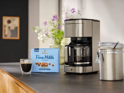 Eine Tasse Kaffee steht neben einer Packung Tchibo Feine Milde Kaffee und einer Kaffeemaschine auf einer Küchentheke.