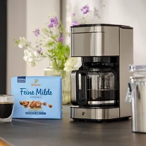 Eine Tasse Kaffee steht neben einer Packung Tchibo Feine Milde Kaffee und einer Kaffeemaschine auf einer Küchentheke.
