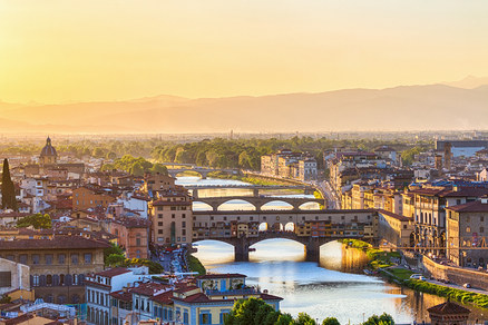 Luftaufnahme von Florenz bei Sonnenuntergang, zeigt die Ponte Vecchio Brücke mit Gebäuden über dem Arno, umgeben von historischen Stadtgebäuden mit roten Dächern und fernen Bergen unter goldenem Himmel.