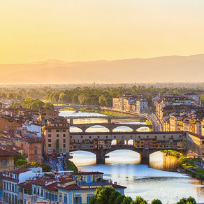 Luftaufnahme von Florenz bei Sonnenuntergang, zeigt die Ponte Vecchio Brücke mit Gebäuden über dem Arno, umgeben von historischen Stadtgebäuden mit roten Dächern und fernen Bergen unter goldenem Himmel.