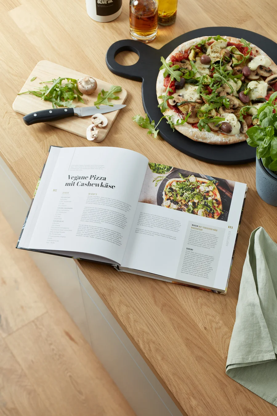 Geöffnetes Buch „Taste of Green – vegan & vegetarisch kochen“ auf Holztisch mit veganer Pizza und Zutaten.