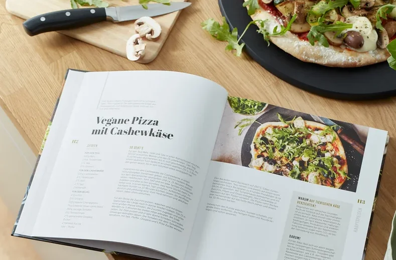 Geöffnetes Buch „Taste of Green – vegan & vegetarisch kochen“ auf Holztisch mit veganer Pizza und Zutaten.