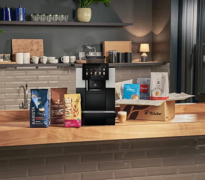 Eine schwarz-silberne Kaffeemaschine steht auf einer Holztheke, umgeben von mehreren Kaffeebohnenbeuteln. Links liegen blaue, braune und gelbe Kaffeebeutel. Rechts befindet sich eine Tchibo-Box mit drei weiteren Kaffeebeuteln und davor ein kleines Glas mit geschichtetem Kaffee.