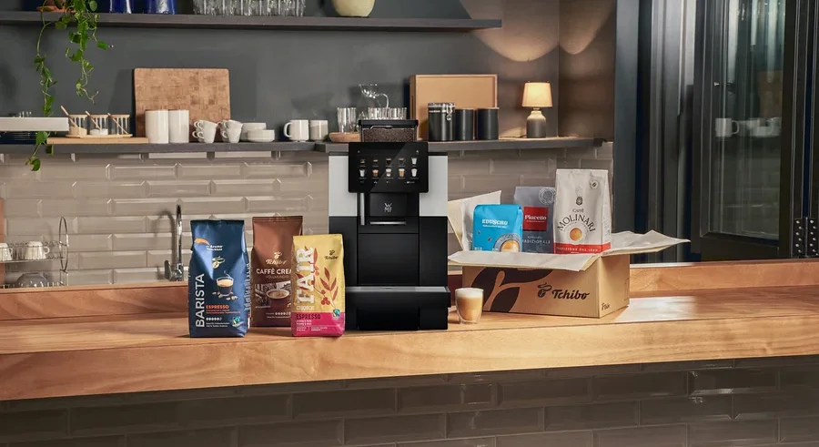 Eine schwarz-silberne Kaffeemaschine steht auf einer Holztheke, umgeben von mehreren Kaffeebohnenbeuteln. Links liegen blaue, braune und gelbe Kaffeebeutel. Rechts befindet sich eine Tchibo-Box mit drei weiteren Kaffeebeuteln und davor ein kleines Glas mit geschichtetem Kaffee.