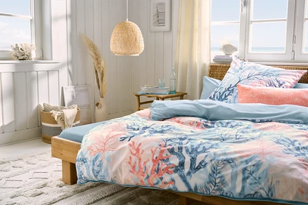 Schlafzimmer mit einem Bett, hellblaues Microfaser-Spannbettlaken in Doppelgröße, Renforcé-Bettwäsche und zwei hellblaue und zwei korallenfarbene Microfaser-Kopfkissenbezüge. Über dem Bett hängt eine Pendelleuchte.