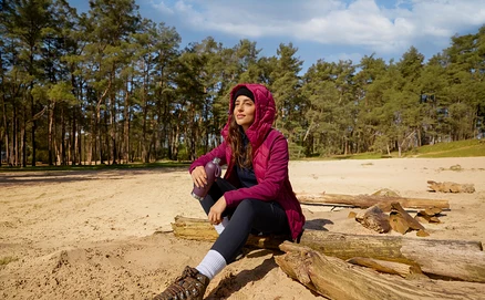 Eine Frau in einer magentafarbenen Jacke sitzt auf einem Baumstamm im Sand, hält eine Sport-Trinkflasche und blickt in den Himmel.