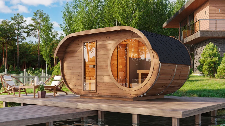 Hölzerne Fasssauna auf einer Holzterrasse über Wasser, mit Gartenmöbeln, umgeben von Bäumen und einem Haus im Hintergrund.