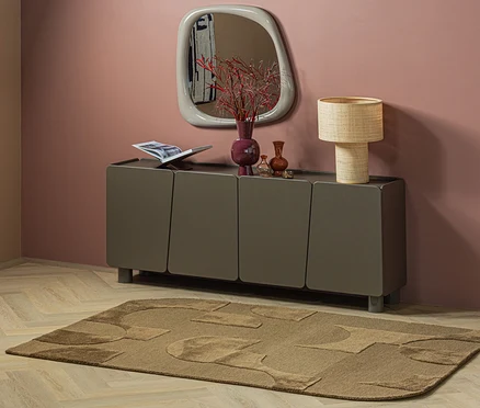 Ein brauner WOOOD Teppich »Livo« mit abstrakten Mustern liegt auf hellem Holzboden vor einem dunkelgrauen Sideboard. Darauf stehen eine offene Zeitschrift, eine lila Vase mit roten Zweigen, kleine braune Vasen und eine geflochtene Tischlampe. Ein unregelmäßig geformter Spiegel hängt an der rosa Wand.