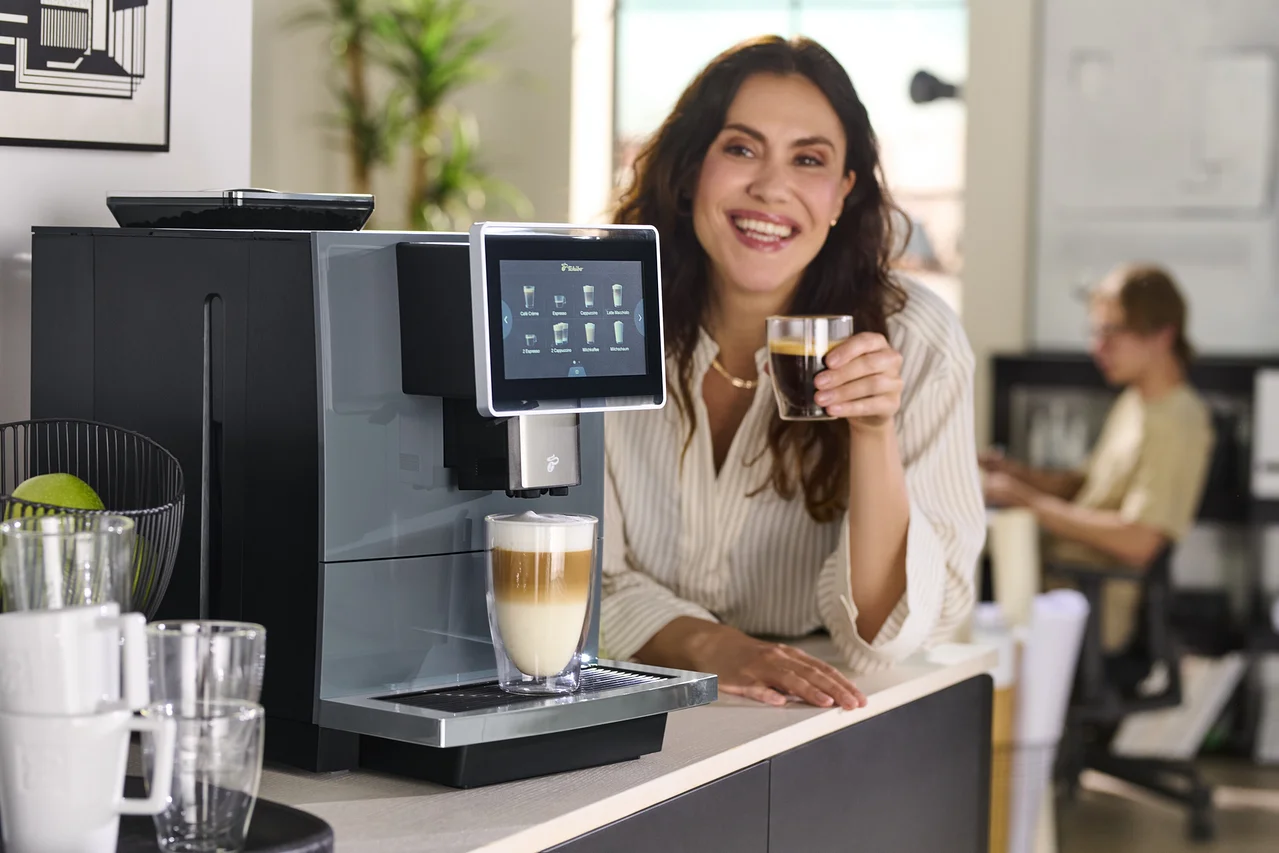Frau hält ein Glas Kaffee neben einer Kaffeemaschine.