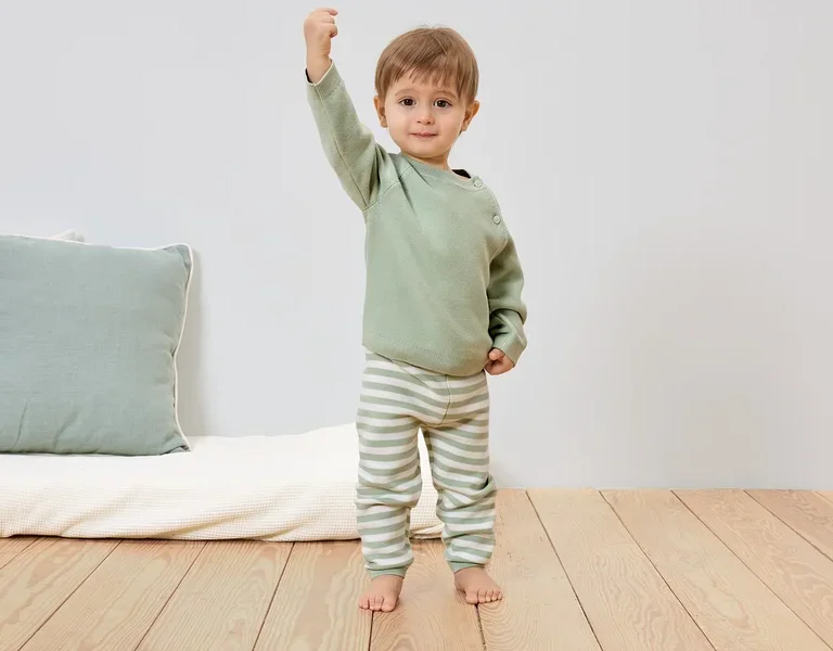 Ein Kleinkind in hellgrünem Pullover und gestreiften Hosen steht barfuß auf einem Holzboden und hebt eine Faust.