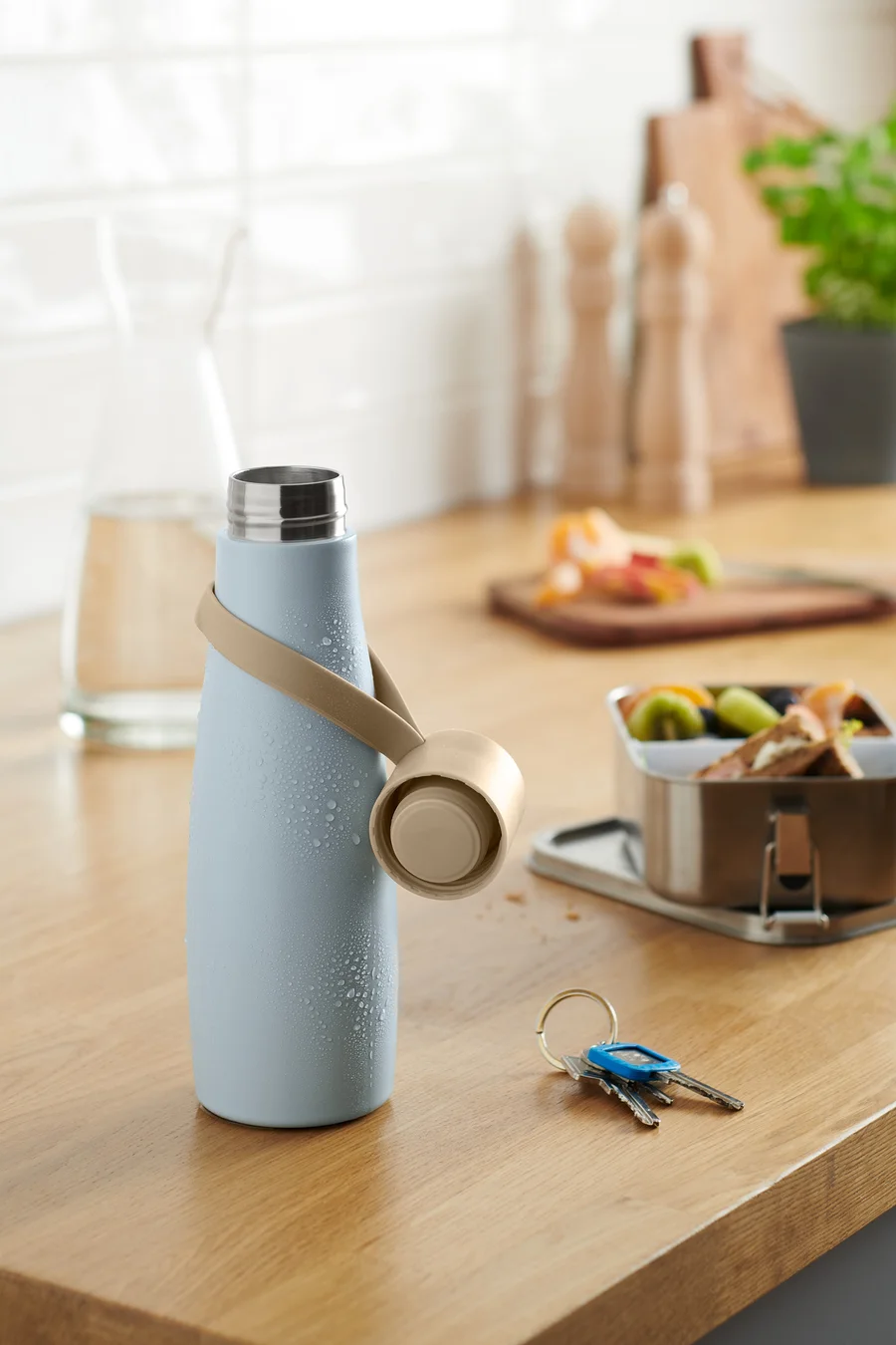 Eine hellblaue To-go-Trinkflasche mit beigem Trageband auf einer hölzernen Küchentheke.