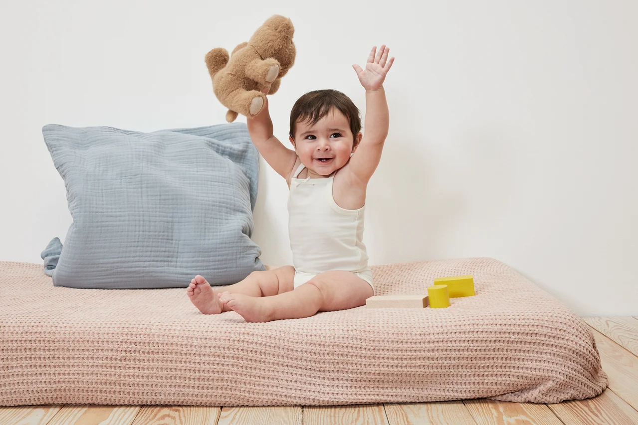 Ein lächelndes Baby sitzt und hält einen Teddybär hoch. Es trägt 5 Baby-Bodys.