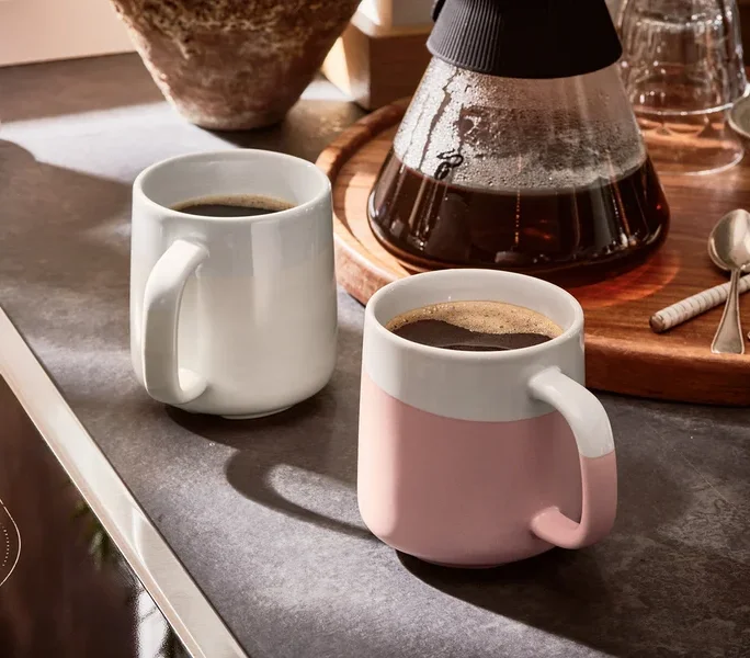 Zwei Kaffeetassen stehen neben einer Kaffeemaschine auf einem Tisch.