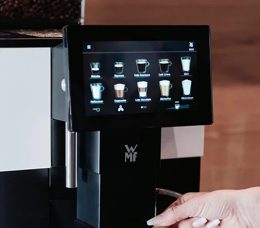 Eine Hand bedient eine schwarze WMF-Kaffeemaschine, um einen klaren Glasbecher zu füllen.