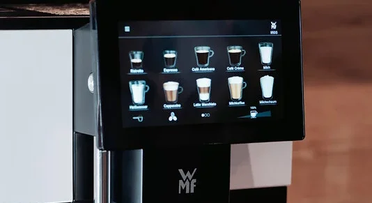 Eine Hand bedient eine schwarze WMF-Kaffeemaschine, um einen klaren Glasbecher zu füllen.