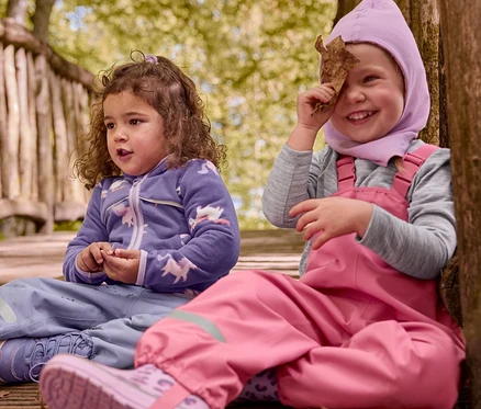 Zwei Kinder sitzen auf einem Holzsteg. Ein Kind trägt lila Thermostiefel und ein lila Einhorn-Oberteil. Das andere Kind trägt pinke Latzhose, pinke Haube und pinke Gummistiefel mit Leo-Muster, hält ein Blatt vors Gesicht.