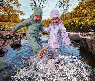 Zwei kleine Kinder in grünen und lila wasserdichten Anzügen planschen im flachen Bach, halten Händchen, mit Herbstbäumen im Hintergrund.