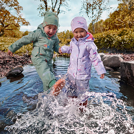 Zwei kleine Kinder in grünen und lila wasserdichten Anzügen planschen im flachen Bach, halten Händchen, mit Herbstbäumen im Hintergrund.