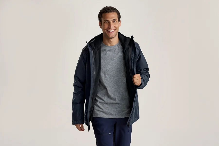 Ein lächelnder Mann trägt eine dunkelblaue Jacke über einem grauen T-Shirt und dunkelblauen Hosen. Er hält die Jacke mit der rechten Hand offen.