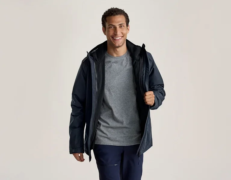 Ein lächelnder Mann trägt eine dunkelblaue Jacke über einem grauen T-Shirt und dunkelblauen Hosen. Er hält die Jacke mit der rechten Hand offen.