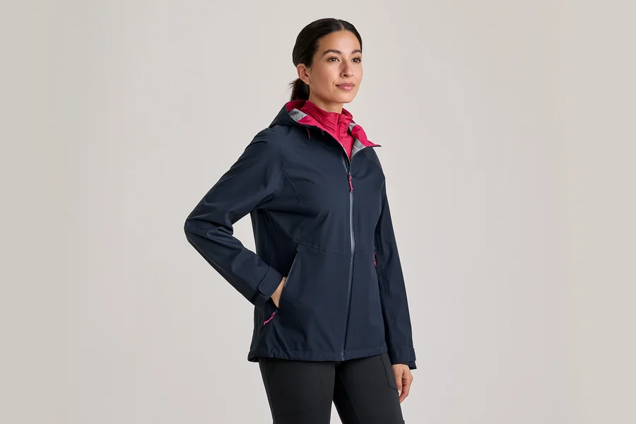 Frau trägt eine dunkelblaue Jacke mit pinkem Innenfutter und schwarzen Hosen, die Hand in der Tasche.