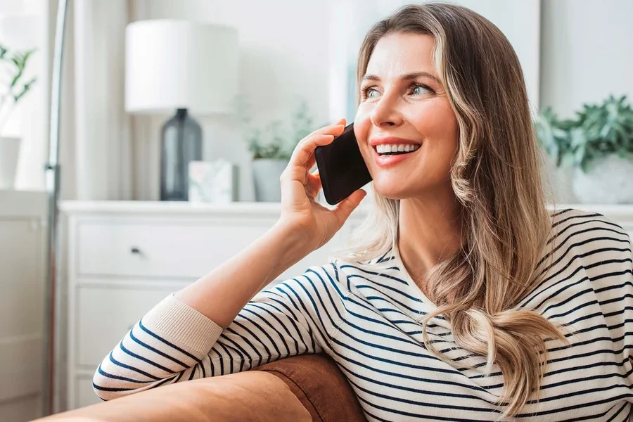 Eine lächelnde Frau telefoniert mit einem Handy, während sie auf einem braunen Sofa im Wohnzimmer sitzt.