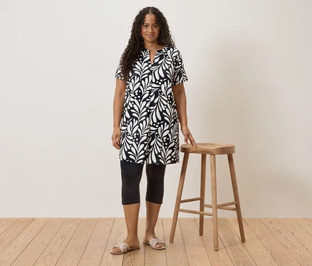 Frau mit lockigem Haar, die ein schwarz-weißes Kleid, Leggings und Sandalen trägt und neben einem Hocker steht.
