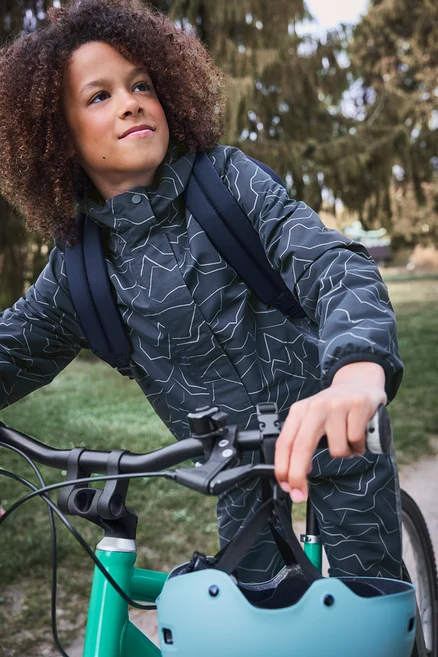 Kind mit lockigem Haar, dunkler gemusterter Jacke und Rucksack steht neben einem grünen Fahrrad, ein hellblauer Helm hängt am Lenker, blickt nach oben.