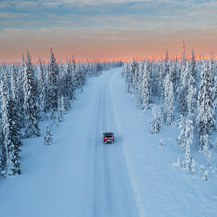 Rotes Auto fährt auf einer verschneiten Straße durch einen Winterwald, mit einem rosa-orangefarbenen Himmel am Horizont.