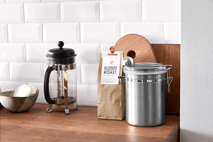 Auf einer Holzplatte stehen eine French Press, eine Schüssel mit Papierfiltern, eine Packung Blonde Roast Kaffee und eine Edelstahldose.