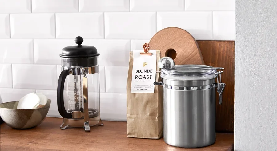 Auf einer Holzplatte stehen eine French Press, eine Schüssel mit Papierfiltern, eine Packung Blonde Roast Kaffee und eine Edelstahldose.