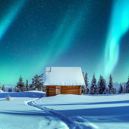 Eine Holzhütte mit schneebedecktem Dach und Eiszapfen steht in einer verschneiten Landschaft mit einem Pfad. Grüne Nordlichter erhellen den sternenklaren Nachthimmel über verschneiten Tannenbäumen.