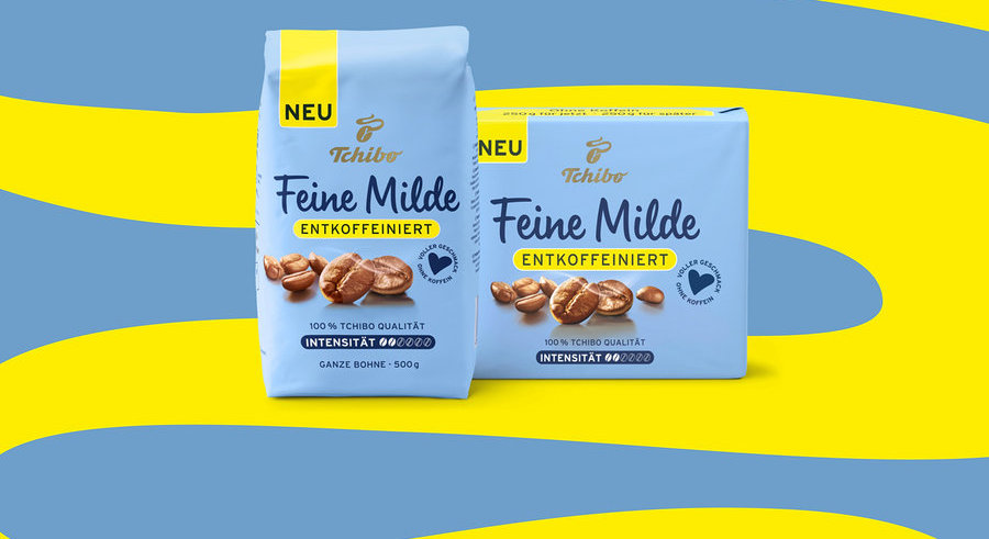 Zwei hellblaue Tchibo Feine Milde entkoffeinierte Kaffeepackungen, eine stehende 500g Bohnenpackung und eine liegende Packung, auf blau-gelbem Hintergrund.