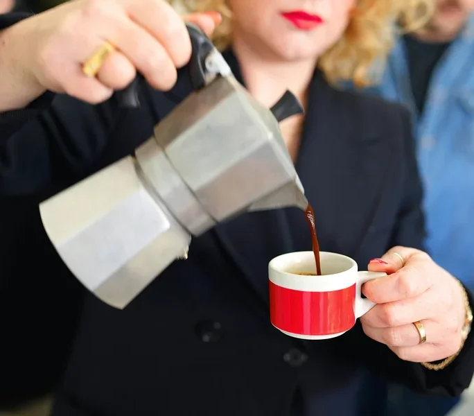 Frau gießt Kaffee aus einer Aluminium-Mokakanne in eine rot-weiße Tasse.