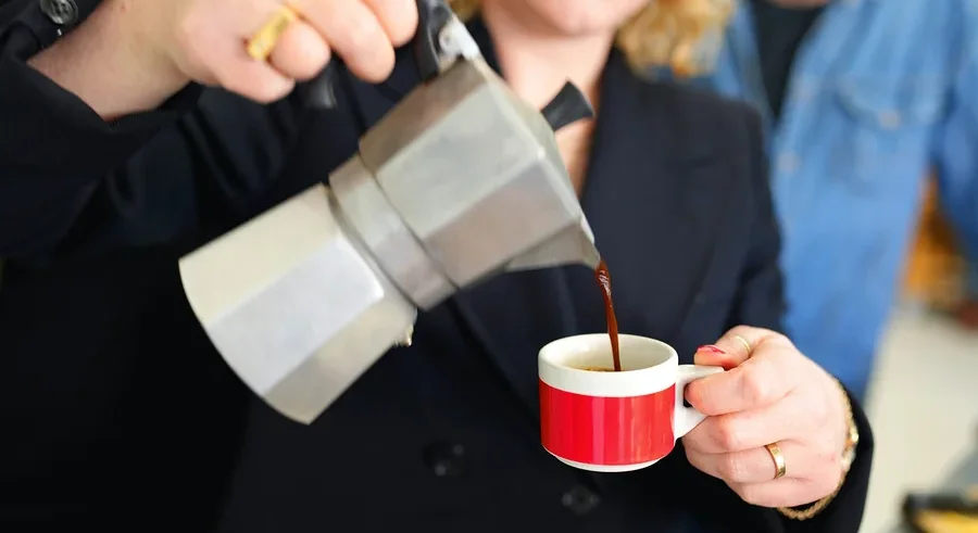 Frau gießt Kaffee aus einer Aluminium-Mokakanne in eine rot-weiße Tasse.