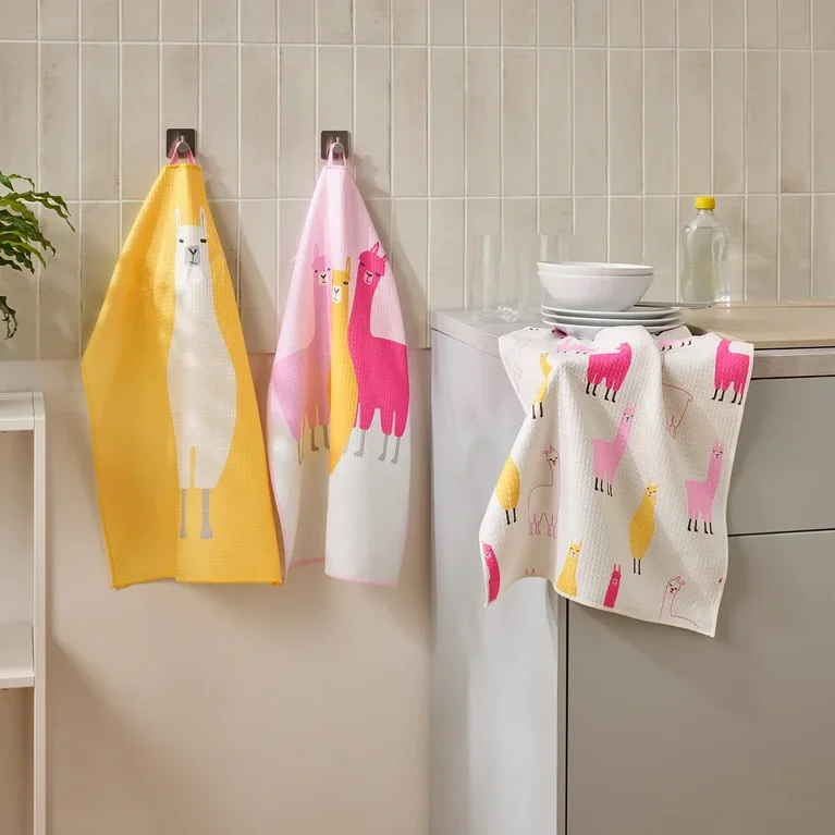 Drei Geschirrtücher mit Lama-Motiven hängen in einer Küche neben einem Waschbecken und einem Regal.