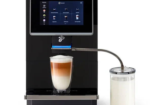 Ein schwarzer Kaffeevollautomat mit Touchscreen bereitet einen Latte Macchiato in einem Glas zu. Ein Milchbehälter ist seitlich angeschlossen.