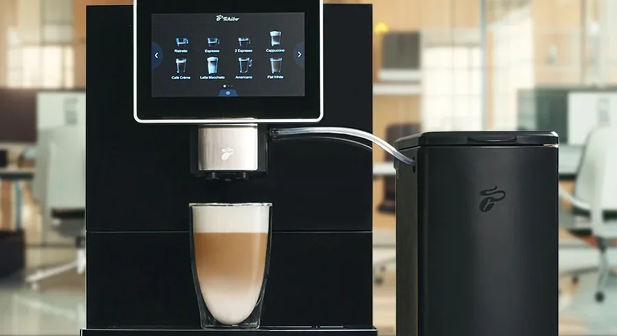 Schwarze Kaffeemaschine mit Touchscreen-Display, das Kaffeeoptionen zeigt, und einem Glas Latte Macchiato darunter. Daneben steht ein schwarzer Milchbehälter.