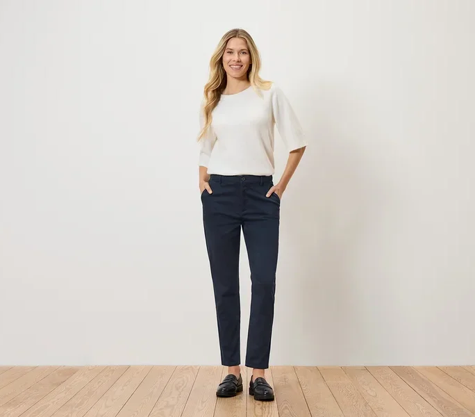 Eine Frau mit blonden, gewellten Haaren steht mit den Händen in den Hosentaschen da. Sie trägt einen weißen Pullover, eine marineblaue Hose und schwarze Schuhe.