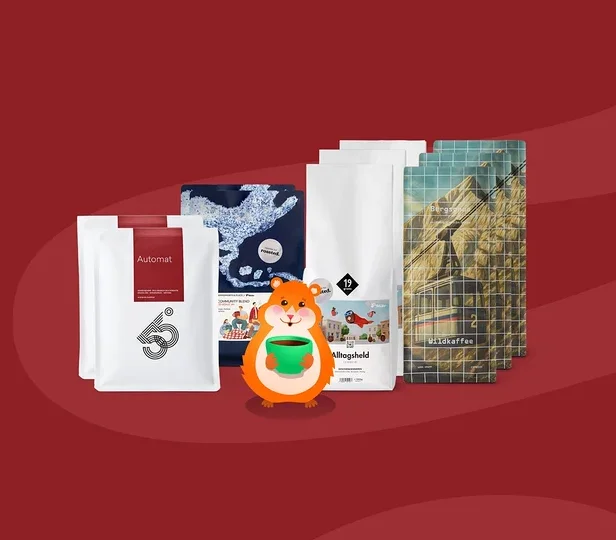 Ein orangefarbener Cartoon-Hamster mit einer grünen Kaffeetasse steht vor fünf Kaffeepackungen. Die Packungen sind weiß mit Aufschriften wie „Automat“ und „Alltagsheld“, eine blaue gemusterte Packung und eine mit Bergmotiv und „Wildkaffee“-Text, auf rotem Hintergrund.