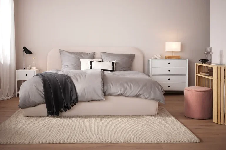 Schlafzimmer mit beigem Bett, grauer Bettwäsche und beigem Teppich. Im Hintergrund weißer Nachttisch und Kommode.